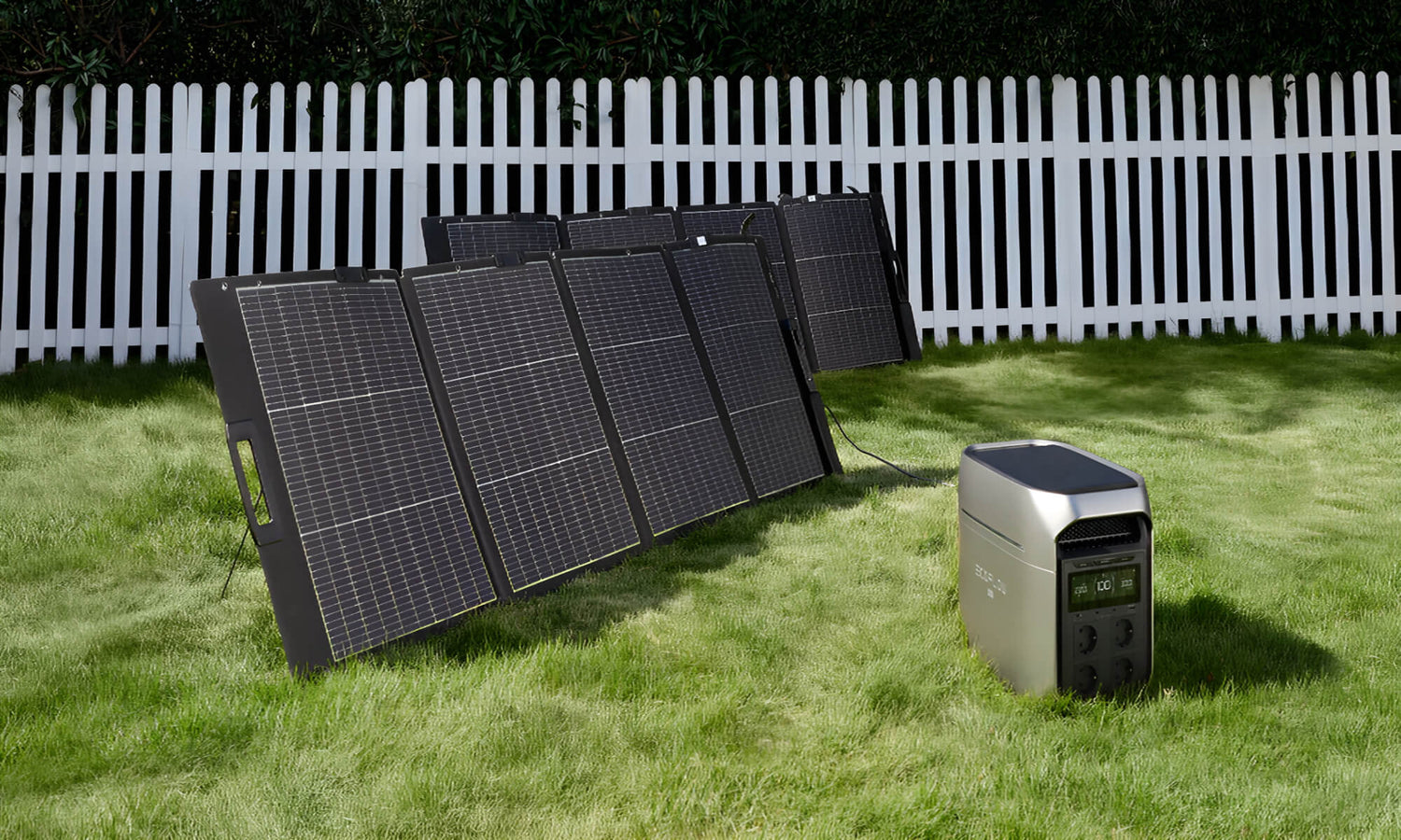 Draagbare zonnepanelen opgesteld op gras in tuin met power station ervoor en witte omheining op achtergrond