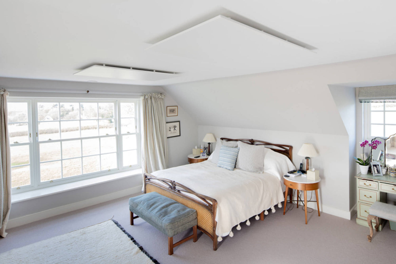 Lichte slaapkamer met een tweepersoonsbed, grote ramen en twee  witte infraroodpanelen op het plafond.